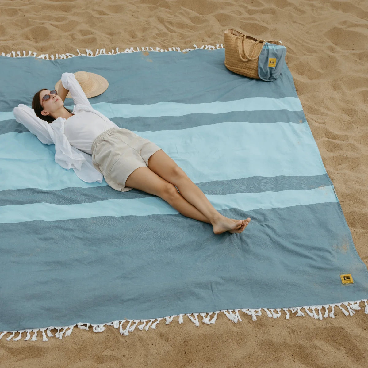 Woman arranging oversized aqua striped blanket on beach sand with white fringe trim