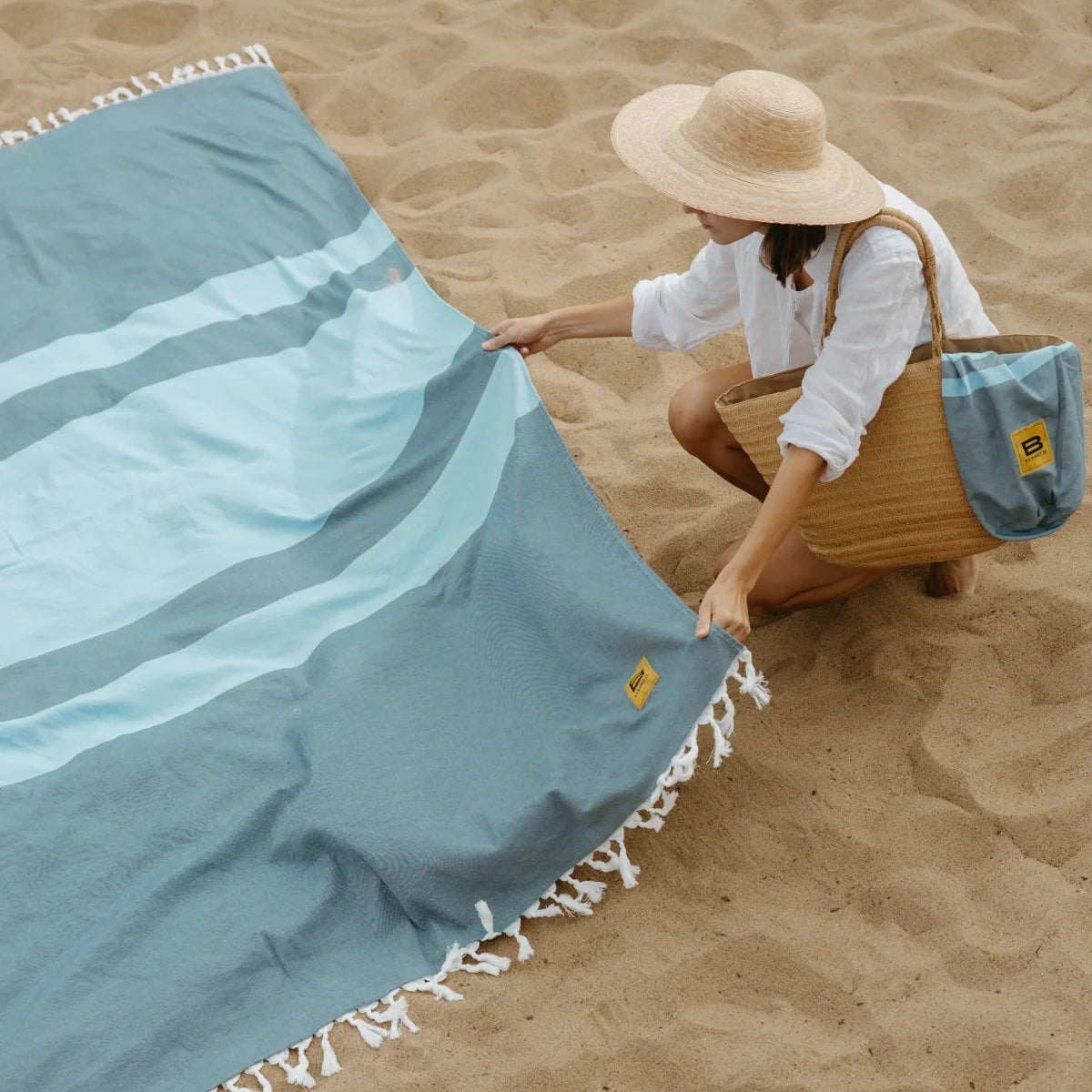 Woman arranging oversized aqua striped blanket with white fringe on beach sand