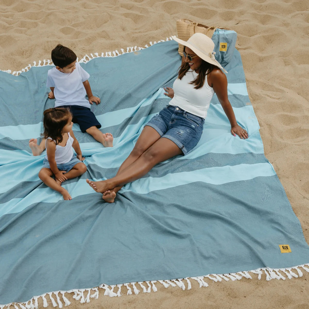 Adult and two children relaxing on oversized blue striped beach blanket with tassel trim in sand