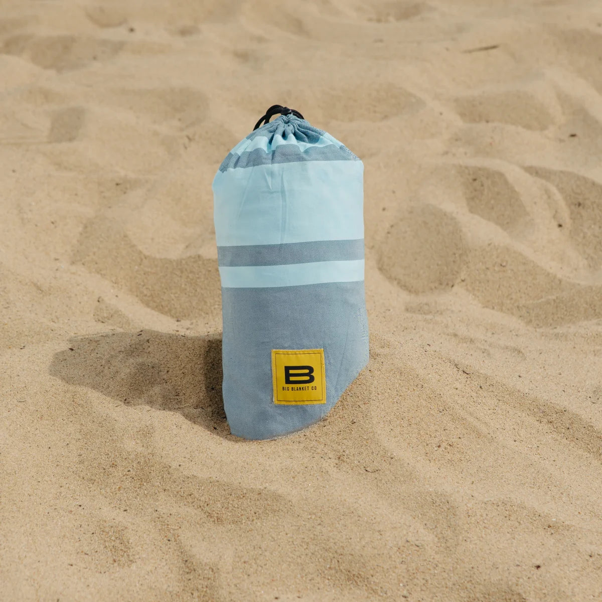 Rolled aqua striped oversized blanket with Big Blanket Co logo on sandy beach