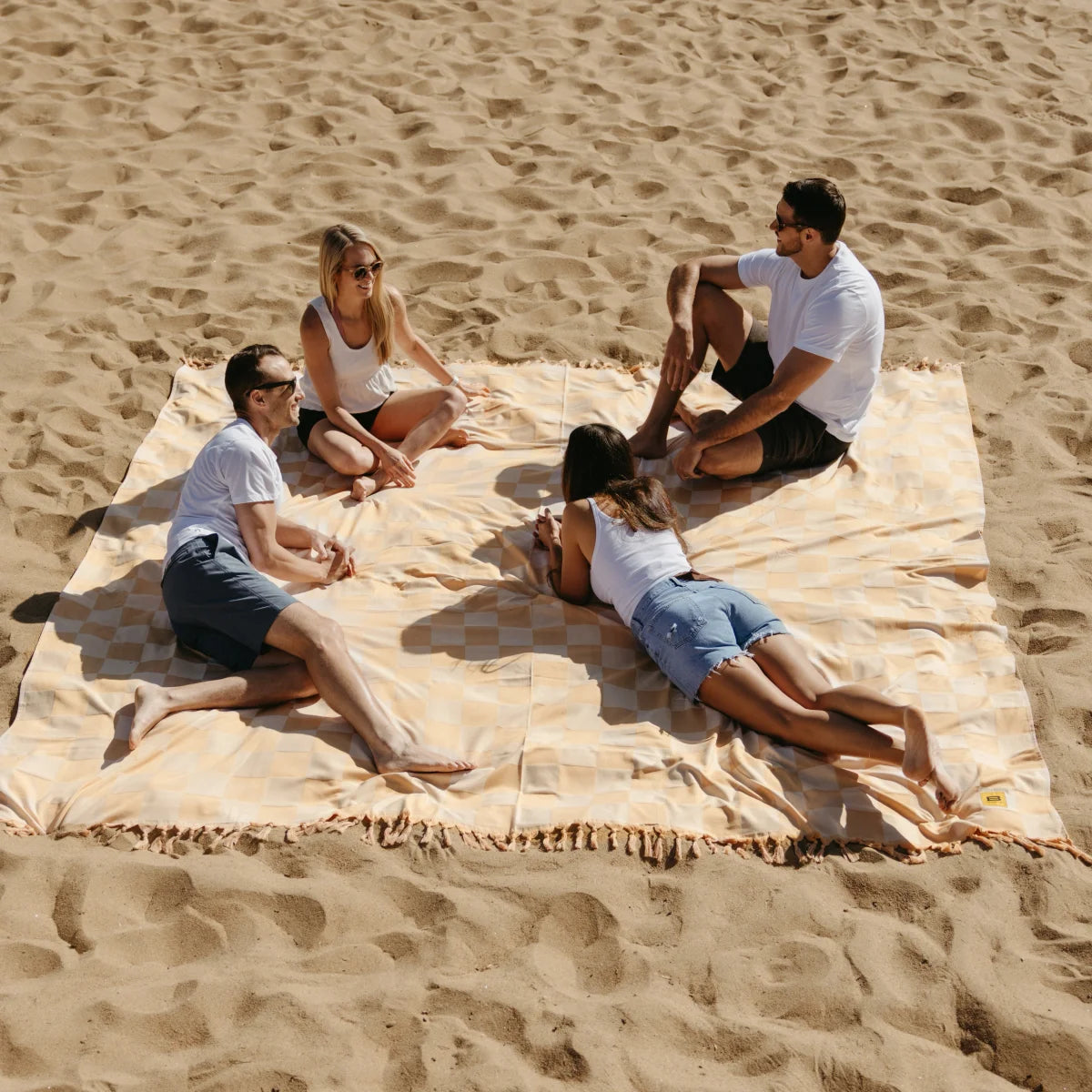 Four people sitting on beige checkered oversized blanket at beach, surrounded by sand