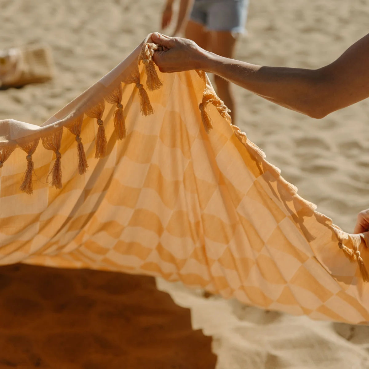 Beige checkered oversized blanket held up on beach, showing soft fabric and fringed edges in sunlight