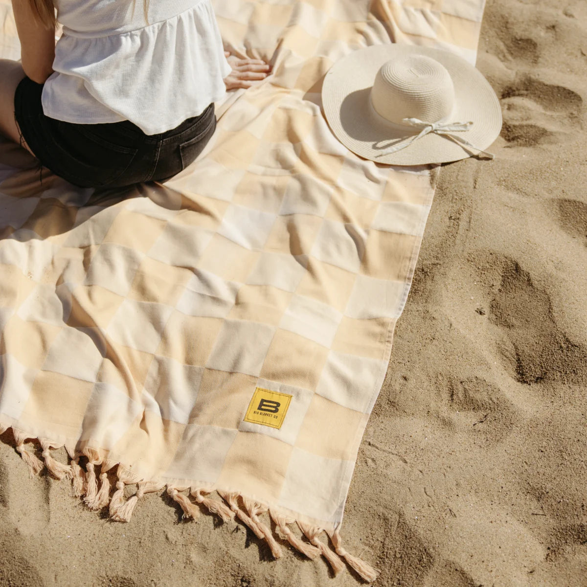 Cream beige oversized blanket with fringe edges spread on sandy beach, person sitting on it wearing white top