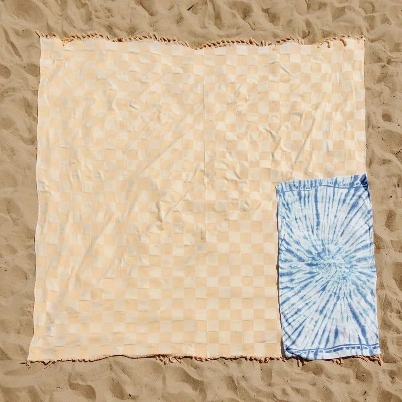 Beige checkered oversized blanket spread on sand with blue tie-dye detail corner, frayed edges