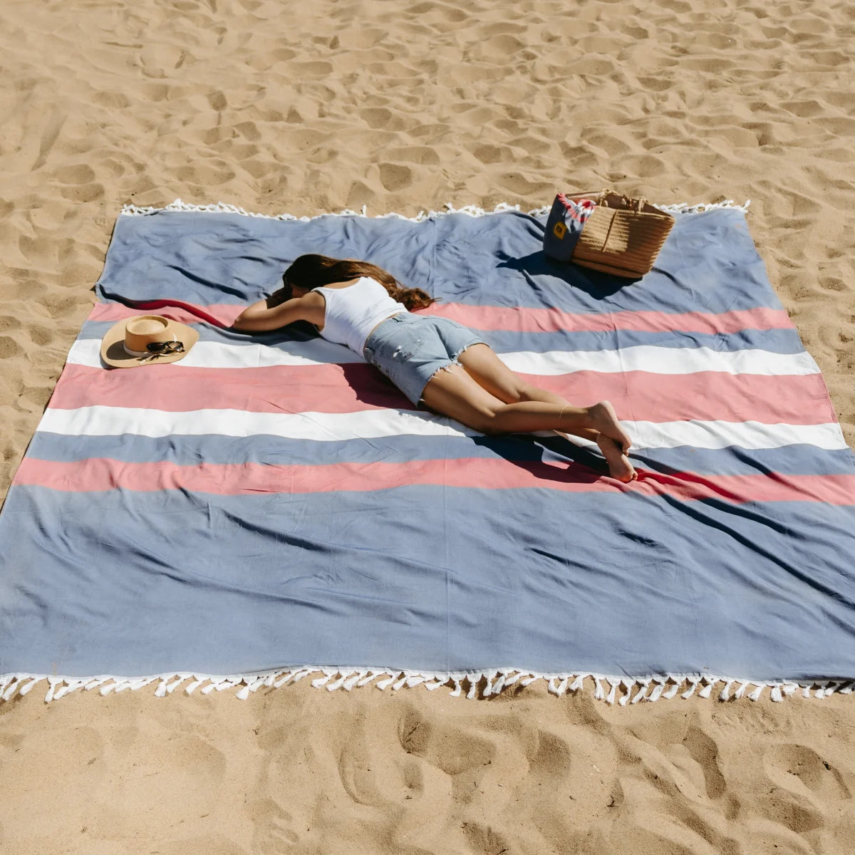 Woman lounging on blue, pink, and white striped oversized beach blanket with tassel fringe in sand