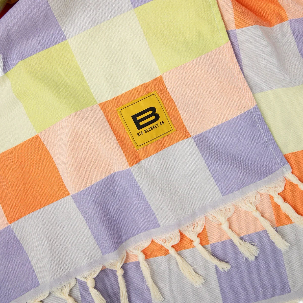 Colorful patchwork oversized blanket with purple, yellow, orange, pink, and cream checkerboard pattern, cream fringe trim, and Big Blanket Co logo label