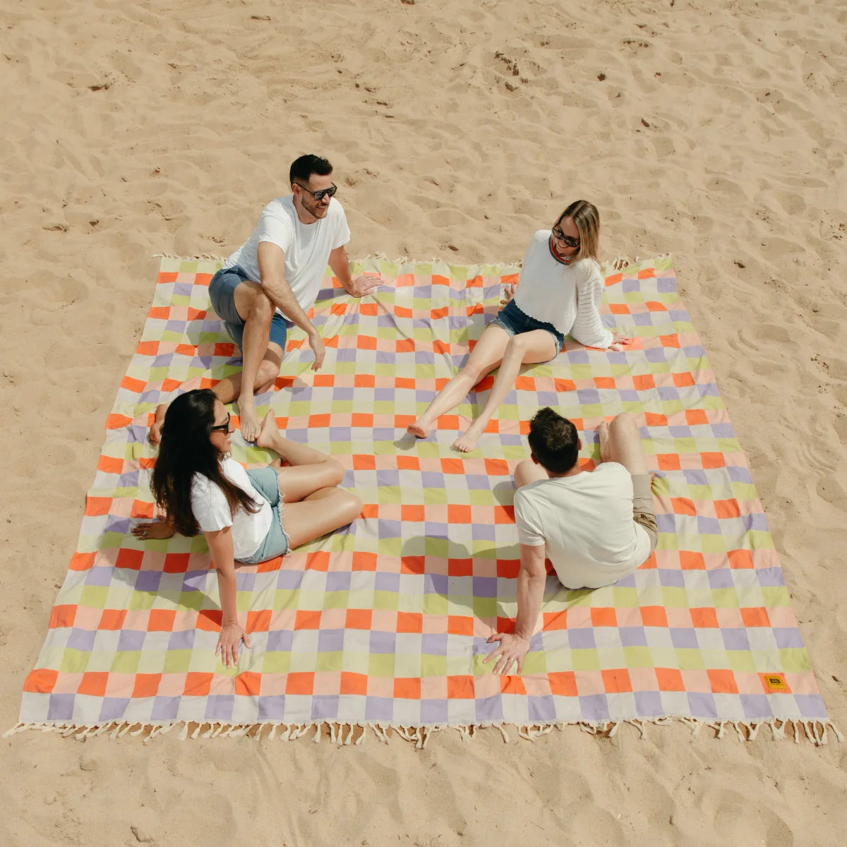 Four people sitting on colorful checkered oversized beach blanket in sand with fringe trim