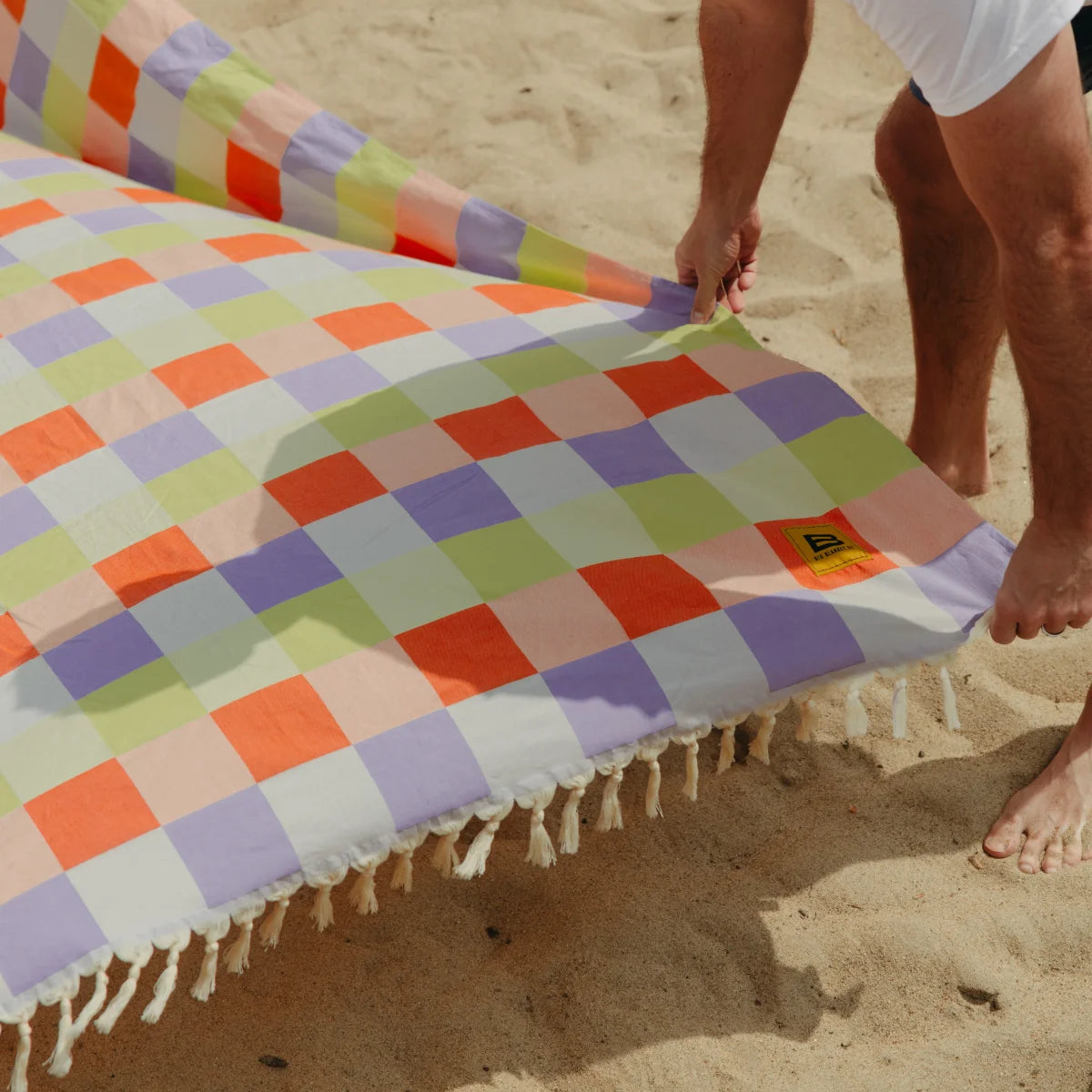 Colorful diamond-patterned oversized blanket spread on beach sand with fringe trim, held by two people barefoot