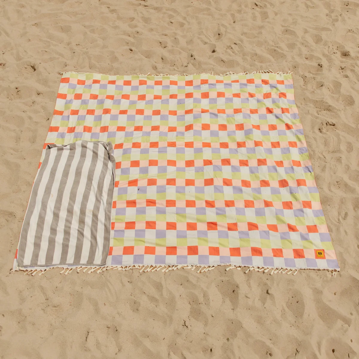 Oversized beach blanket with bright multicolored checkerboard pattern in orange, yellow, lavender and cream, displayed on sand with striped side visible.