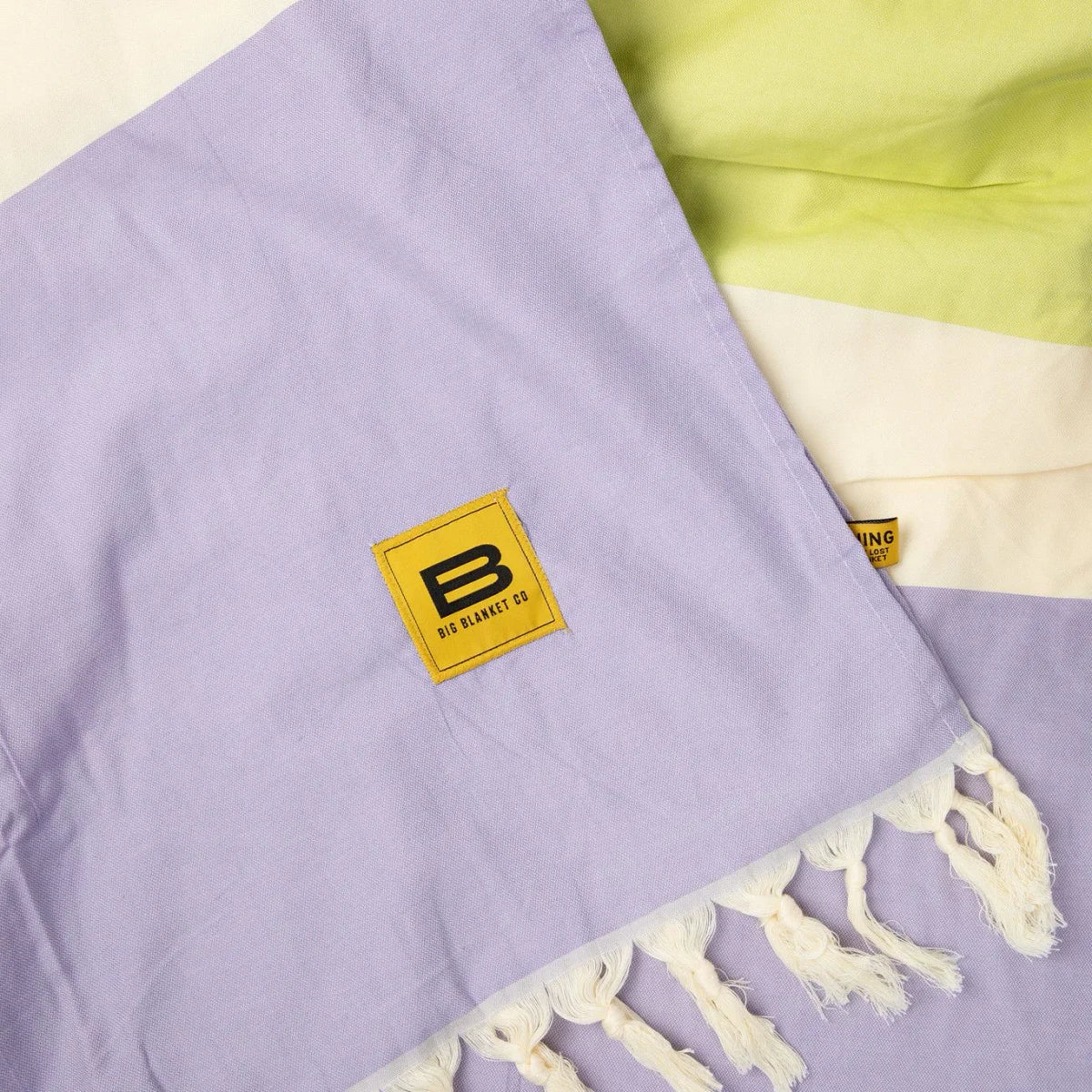 Lavender oversized blanket with fringe trim and yellow Big Blanket Co logo tag, displayed flat with lime stripe detail
