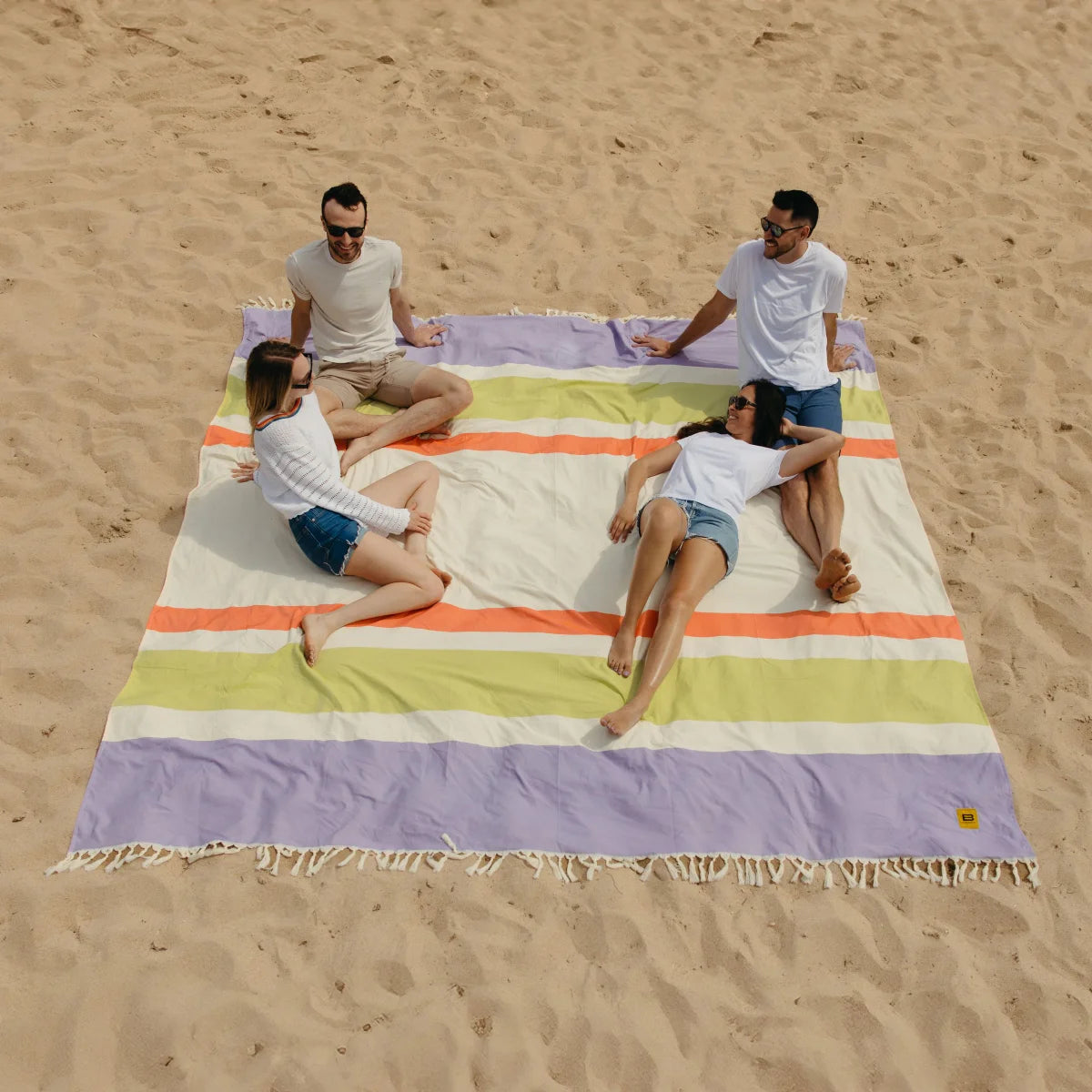 Four people relaxing on colorful striped oversized beach blanket in sand with purple, green, orange, and cream horizontal stripes