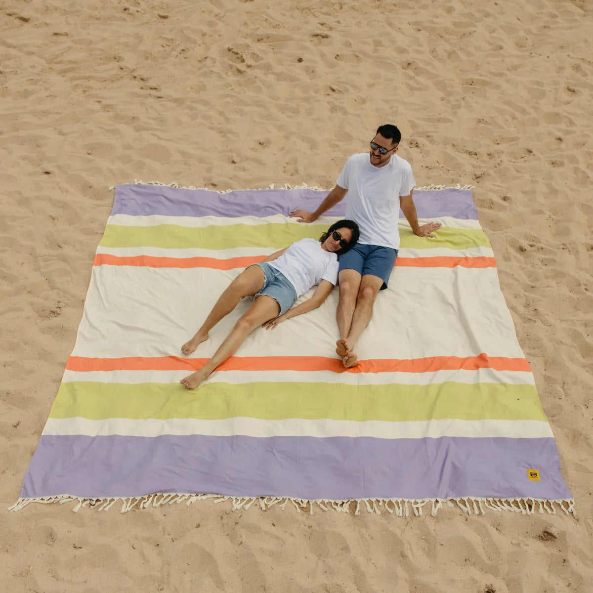 Couple relaxing on oversized striped blanket with purple, yellow, orange, and cream stripes on sandy beach