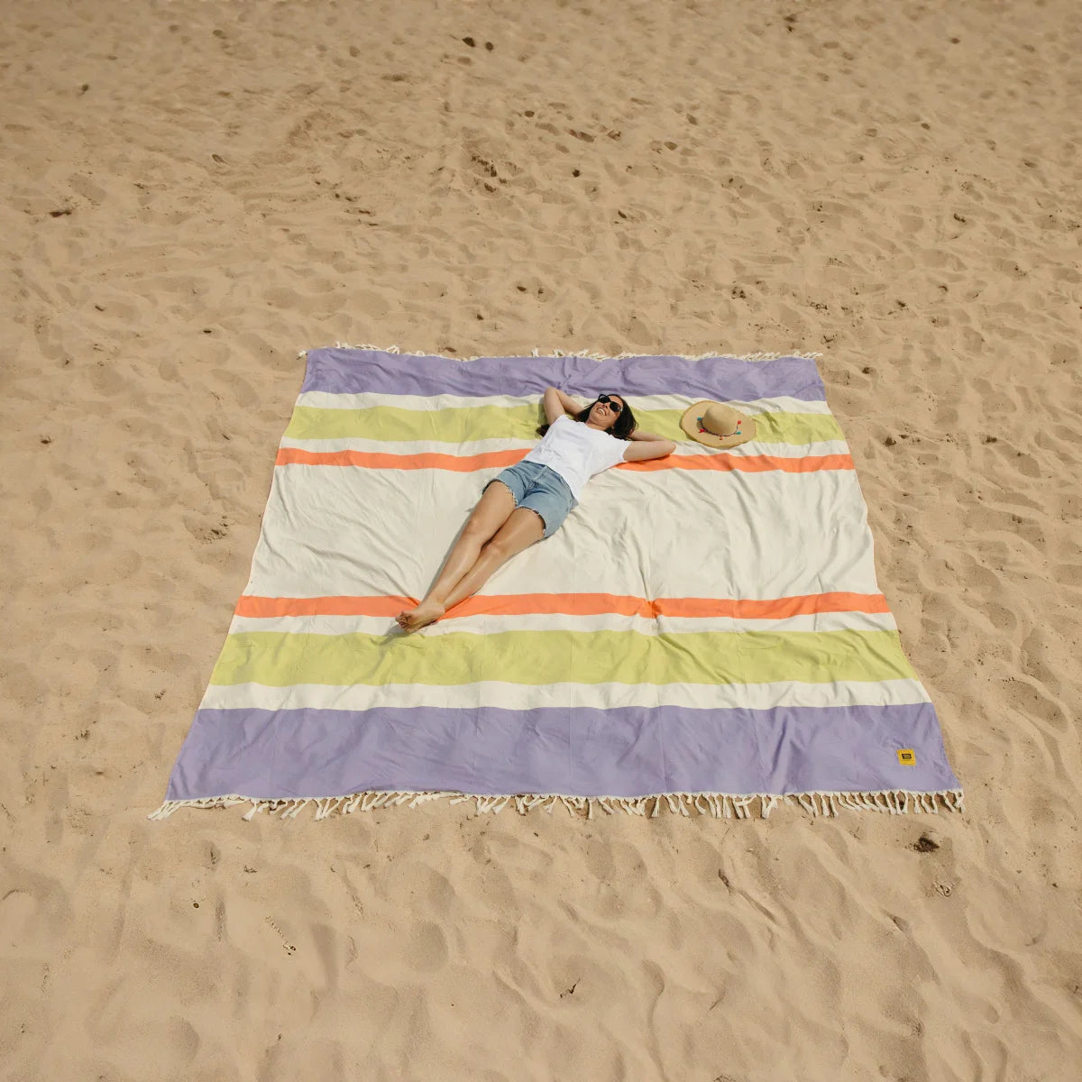 Oversized striped beach blanket in purple, yellow, orange and cream with fringe edges, person lying on sand