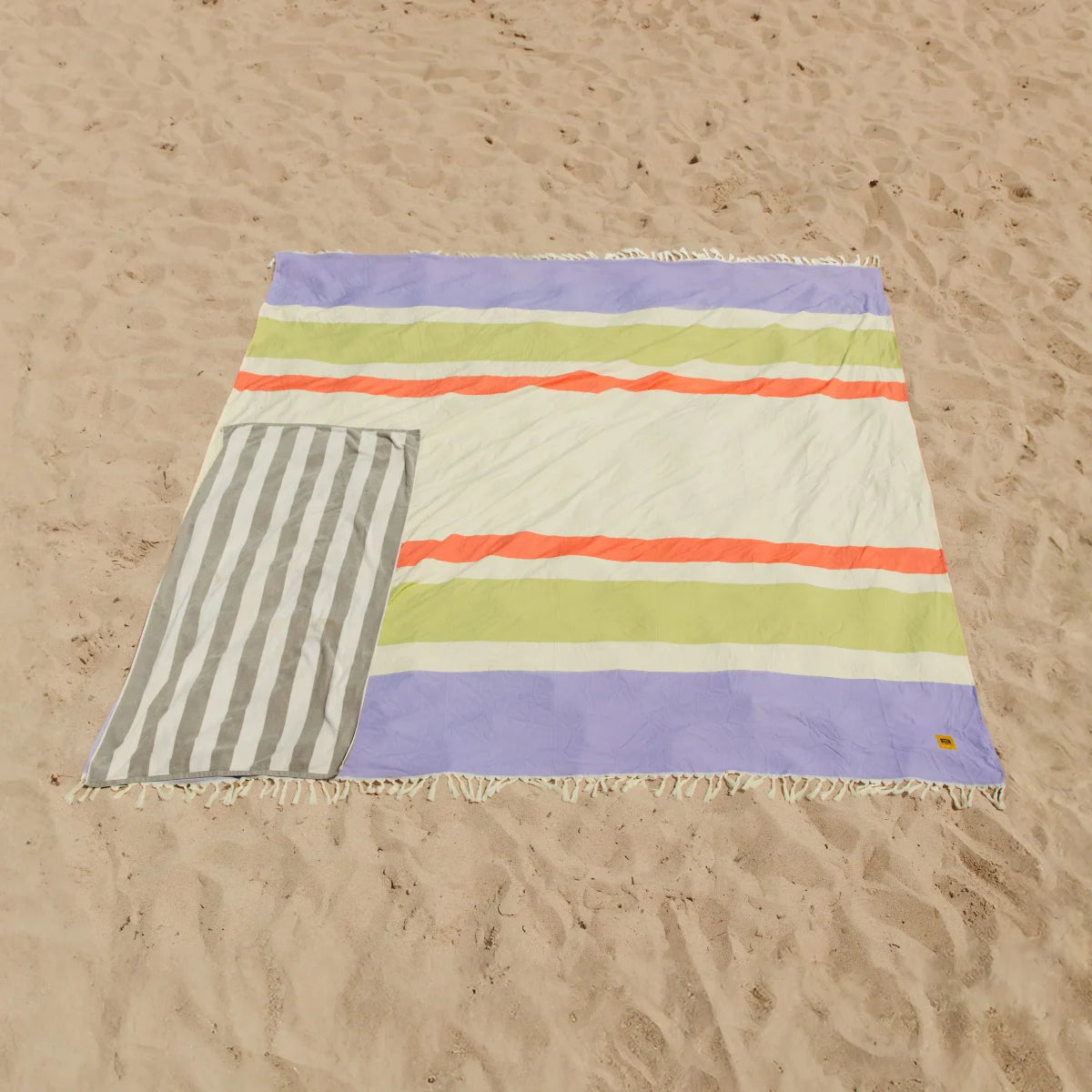 Colorful striped oversized beach blanket with purple, lime, coral, and cream stripes on sand