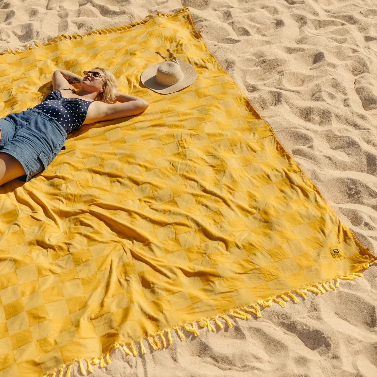 Woman relaxing on golden checkered oversized beach blanket in sand with hat and sunglasses