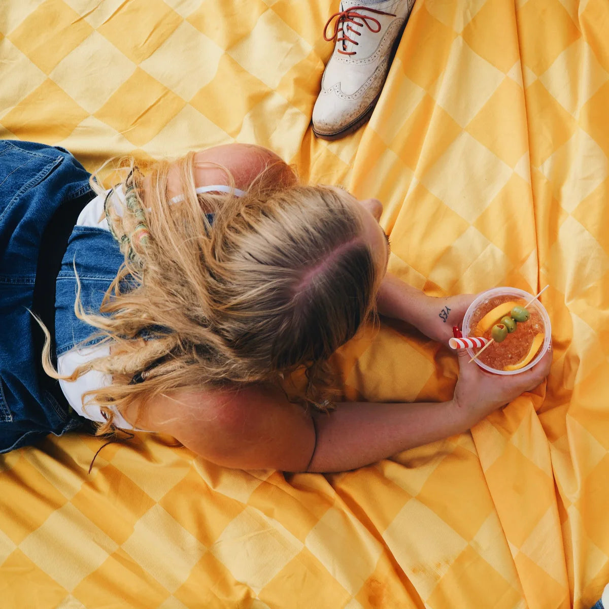 Overhead view of person relaxing on golden checkered oversized blanket holding drink with feet up