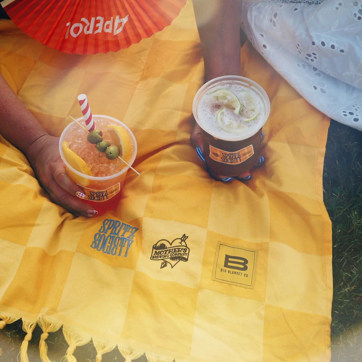 Golden checkered Big Beachy blanket with beverages and red fan, outdoor picnic setting