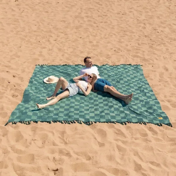 Couple relaxing on green checkered oversized beach blanket in sand, woman in white hat and man in sunglasses