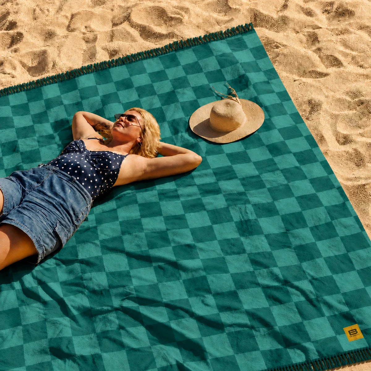 Woman relaxing on teal checkered oversized beach blanket in sand with hat and sunglasses