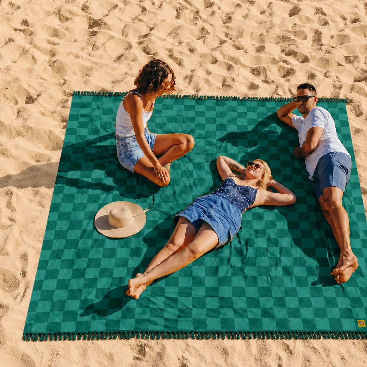 Teal checkered oversized blanket on beach sand with three people lounging and socializing