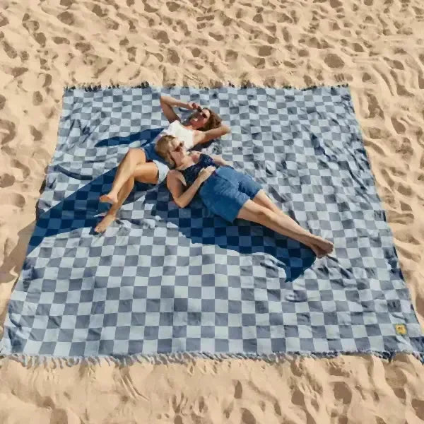 Blue checkered oversized blanket on sand with two people relaxing, beach setting