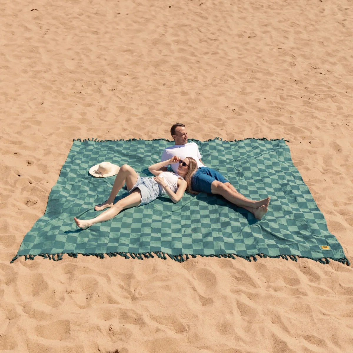 Two people lounging on large green checkered oversized blanket on sandy beach