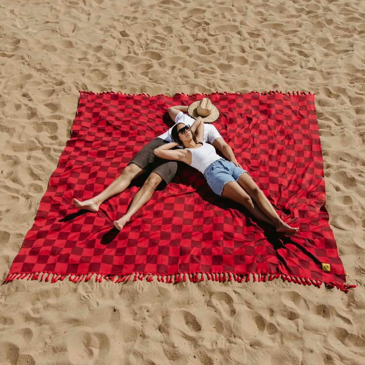 Couple relaxing on red checkered oversized beach blanket in sand