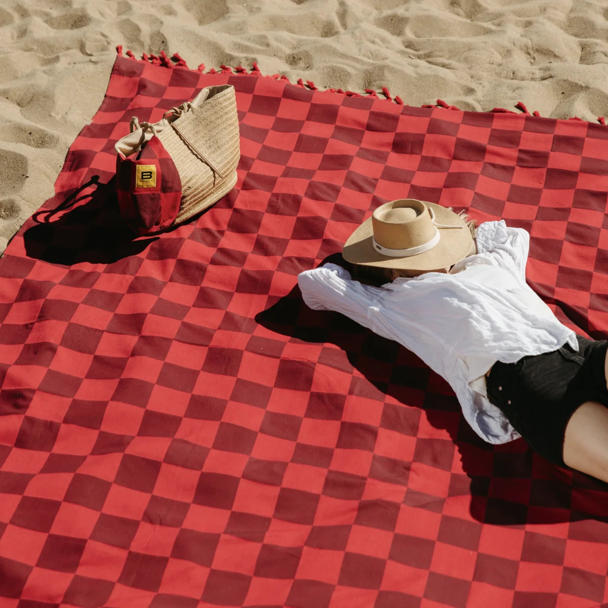 Red checkered oversized blanket on sand beach with person relaxing, hat, and beach accessories