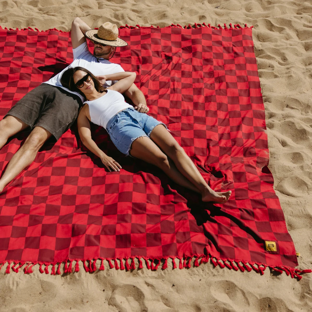 Couple relaxing on red checkered oversized beach blanket in sand with fringe trim