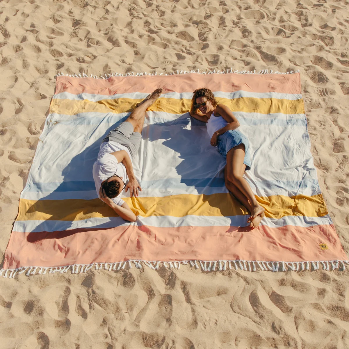Oversized Big Beachy blanket with sunrise stripe pattern in pink, white, yellow, and blue stripes, laid flat on sand with two people relaxing on it.