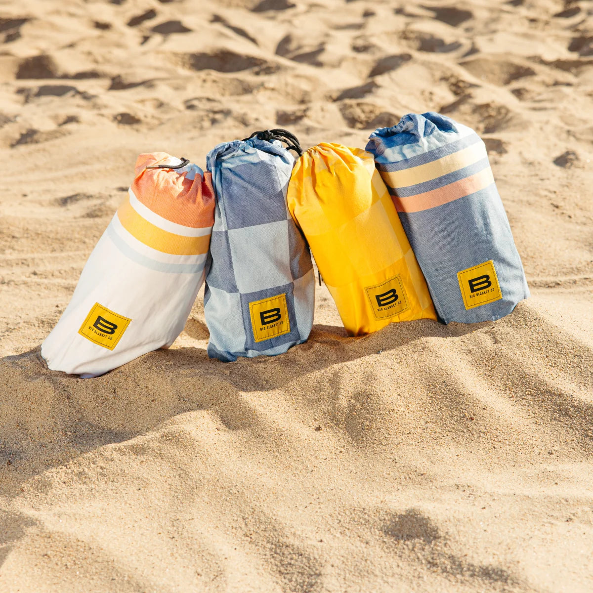 Four rolled Big Blanket Co oversized blankets on beach sand in white, blue, yellow, and striped patterns with yellow logo labels