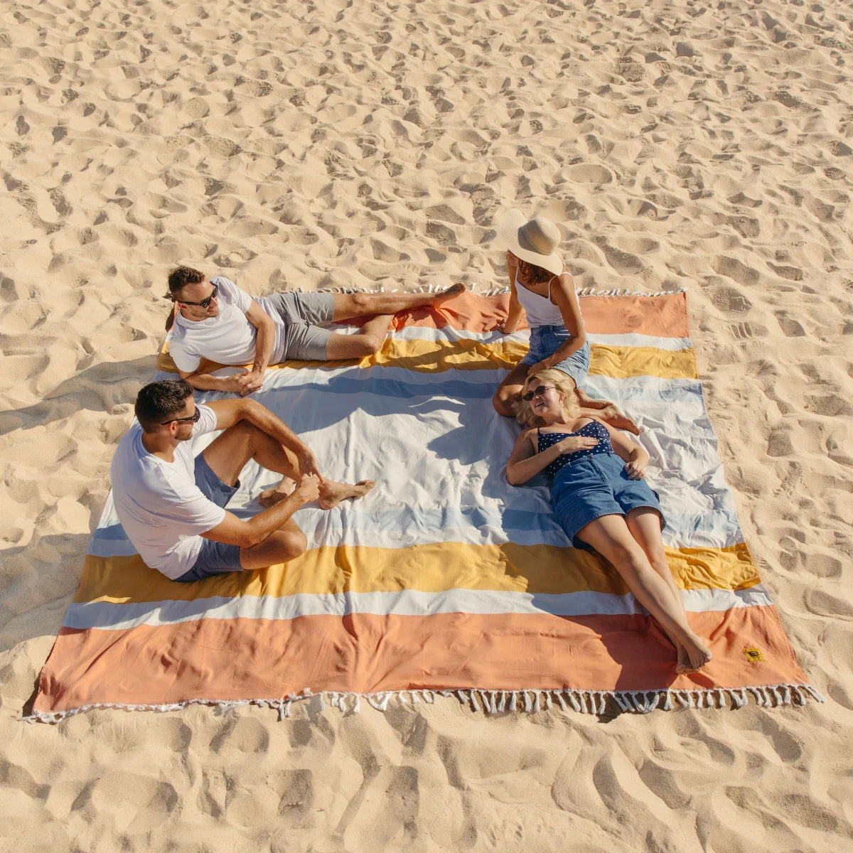 Family of four relaxing on striped oversized beach blanket in sand with yellow, orange, and white horizontal stripes