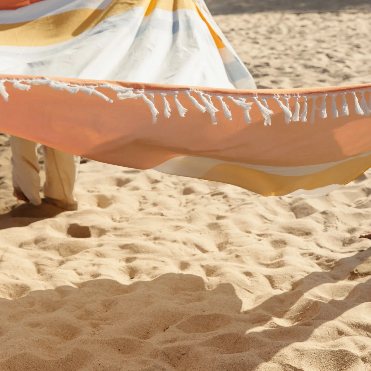 Oversized beach blanket in orange, white, and gold stripes with fringe edge spread on sandy beach