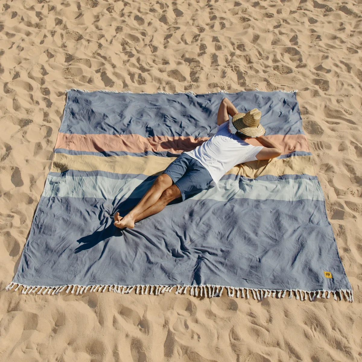 Man relaxing on blue and tan striped oversized beach blanket in sand wearing white shirt and straw hat