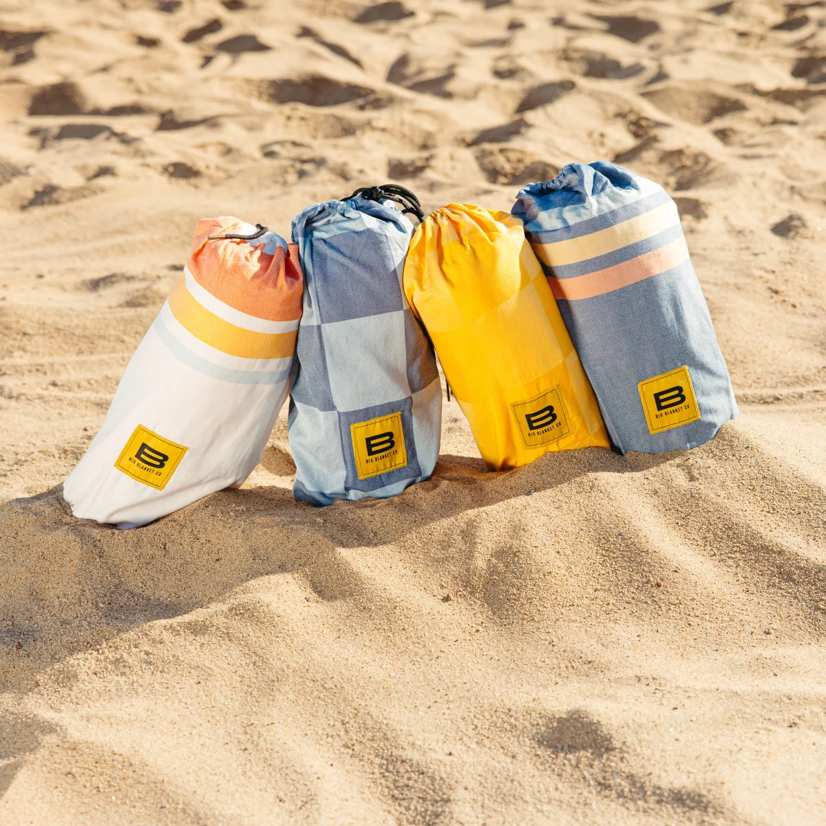 Four rolled Big Beachy blankets in coral, blue, yellow, and striped patterns on sandy beach