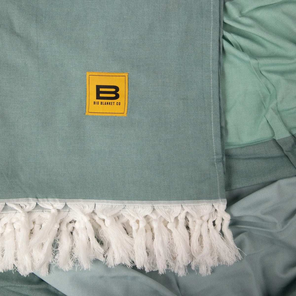 Sage green oversized blanket with white fringe edge and Big Blanket Co logo label detail