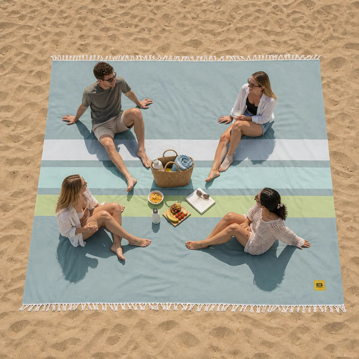 Four friends sitting on striped oversized beach blanket with food and drinks on sand