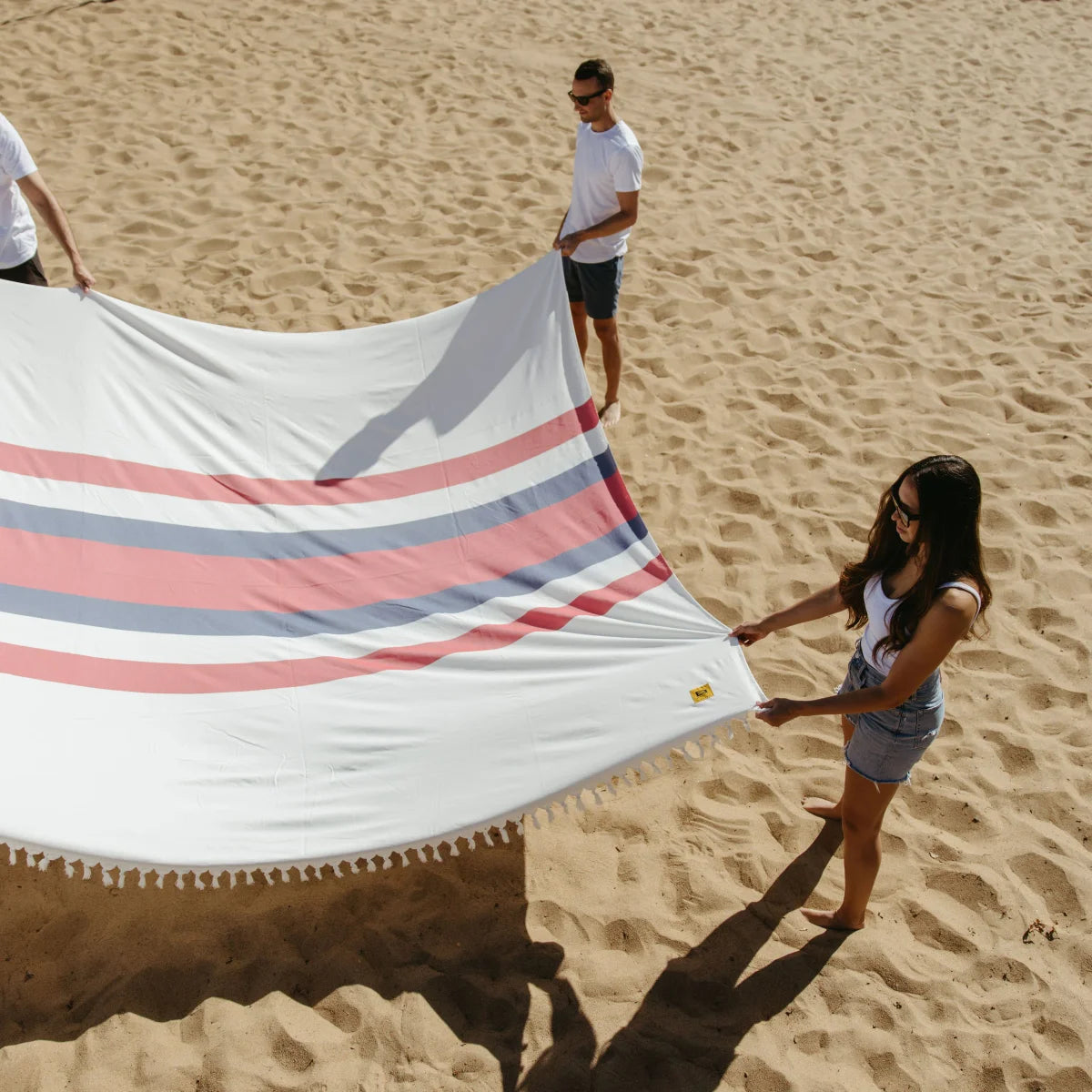 Two people holding oversized white blanket with pink and blue stripes on sandy beach