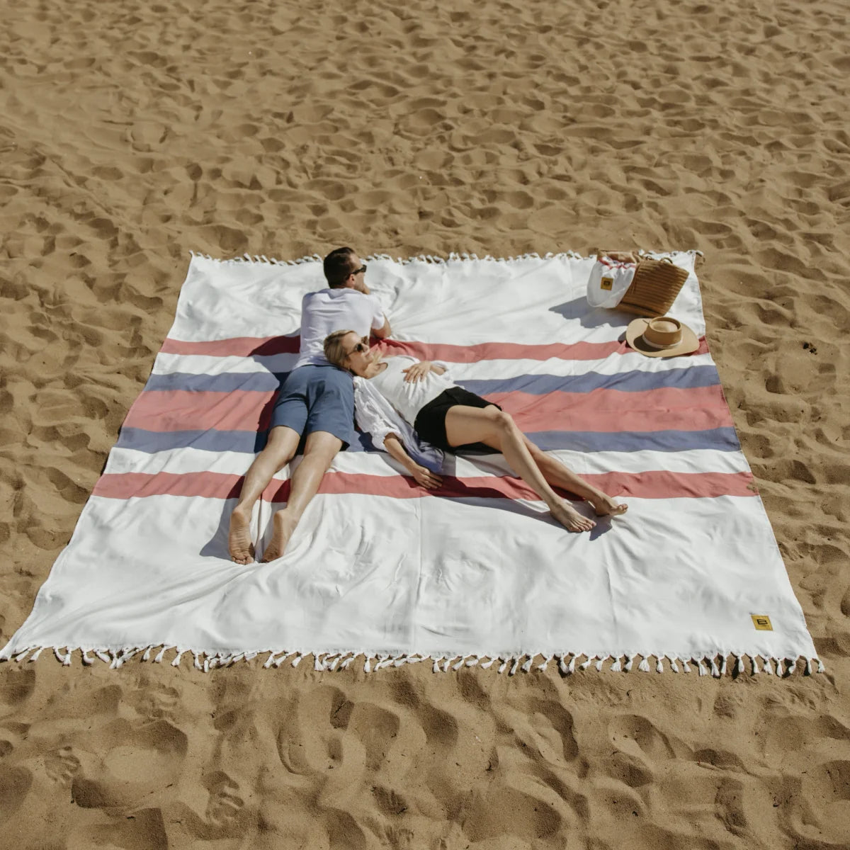 Couple relaxing on oversized white striped beach blanket in sand with hat and book nearby