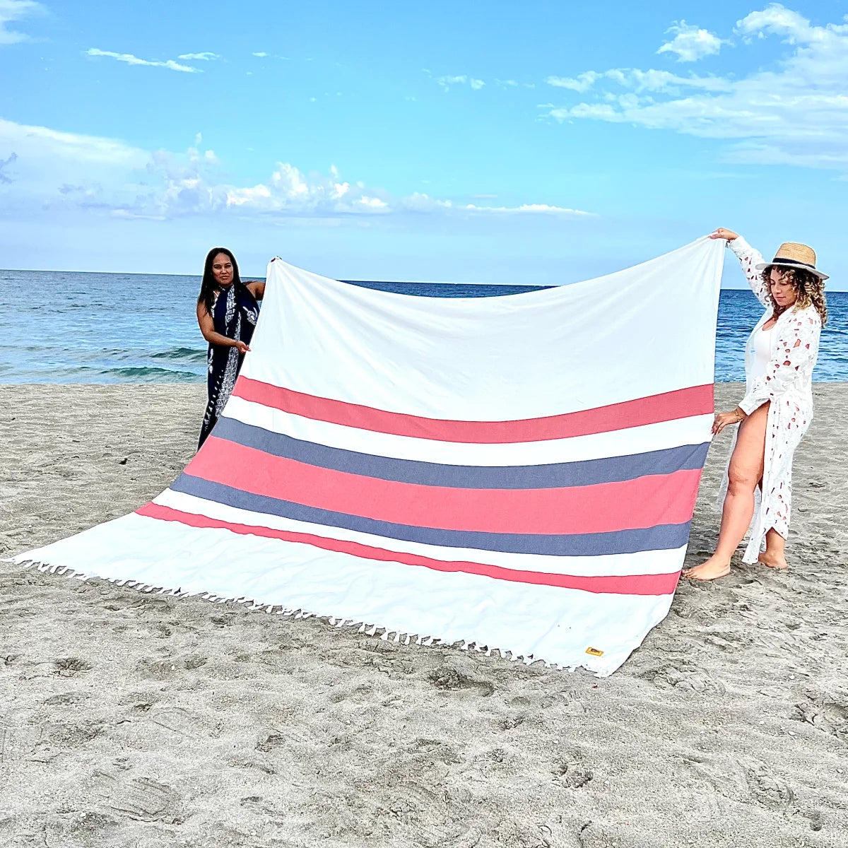 Oversized striped blanket in white, coral, and navy held by two women on beach with ocean backdrop