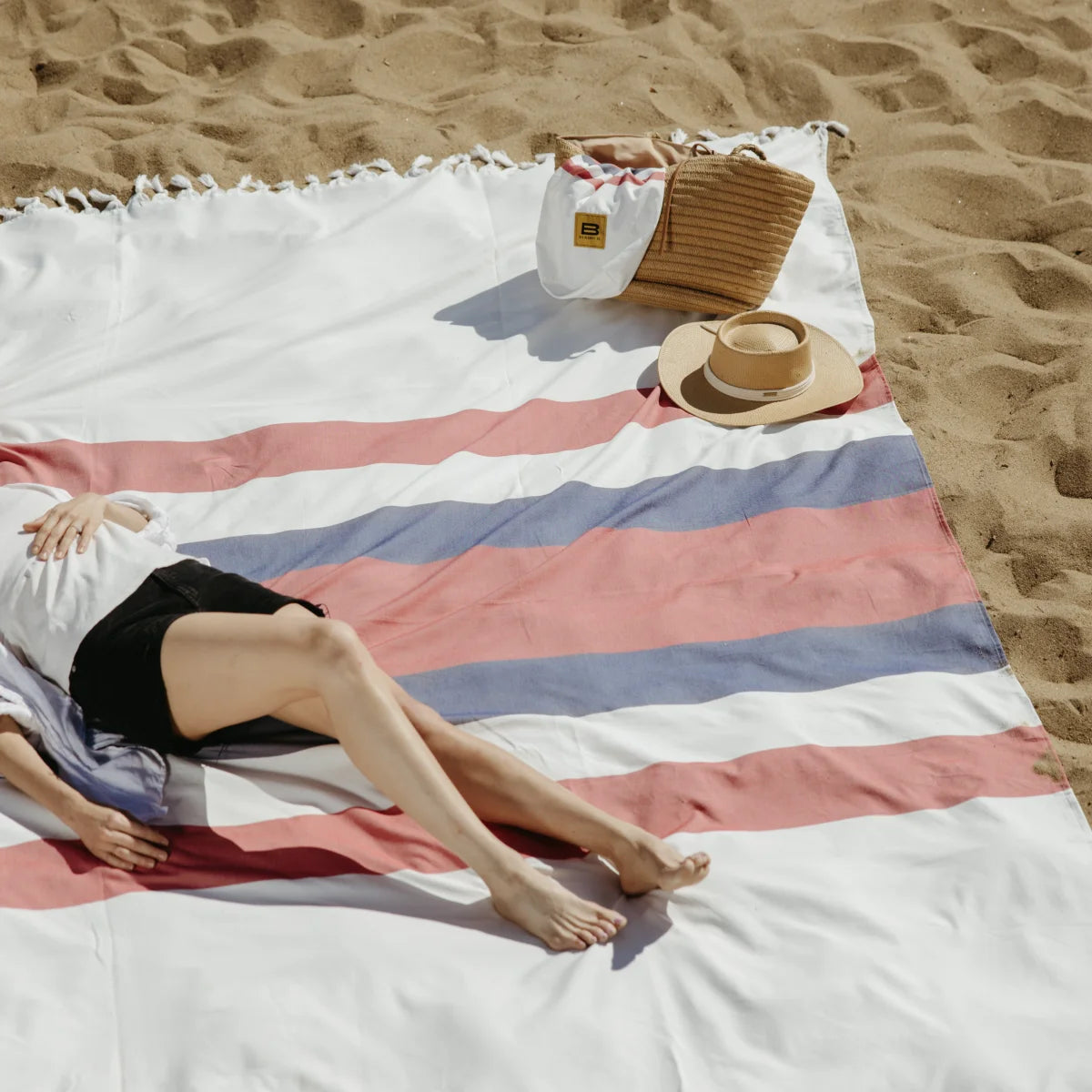 Woman relaxing on striped Big Beachy blanket at beach with hat and beach bag nearby