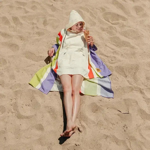Woman wearing cream Big Beachy Poncho with bright rainbow stripes lying on sand beach