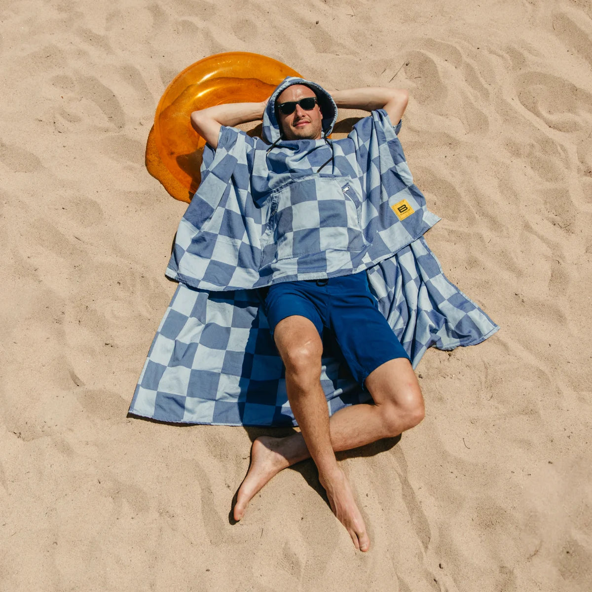 Man relaxing on sandy beach wearing blue and white checkered Big Beachy poncho with hood