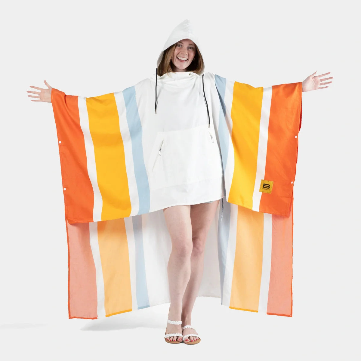 Woman wearing oversized striped poncho blanket in orange, yellow, and blue with white hood, arms spread wide