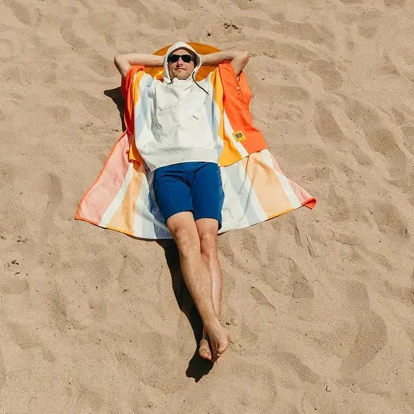 Person lying on sand wearing Big Beachy Poncho in sunrise stripe pattern with orange, pink, and cream colors