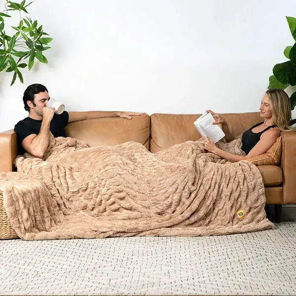 Couple on tan leather couch sharing oversized beige quilted blanket, one reading while relaxed together