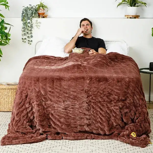 Man sitting in bed under oversized mocha velvet blanket, eating snacks surrounded by pillows and potted plants