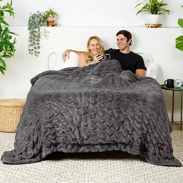 Couple snuggled under charcoal gray oversized blanket with textured quilted pattern on bed with white headboard