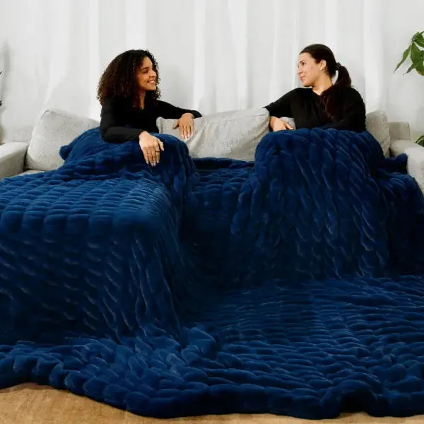 Two women sitting on couch wrapped in deep blue quilted oversized blanket, textured fabric draping to floor