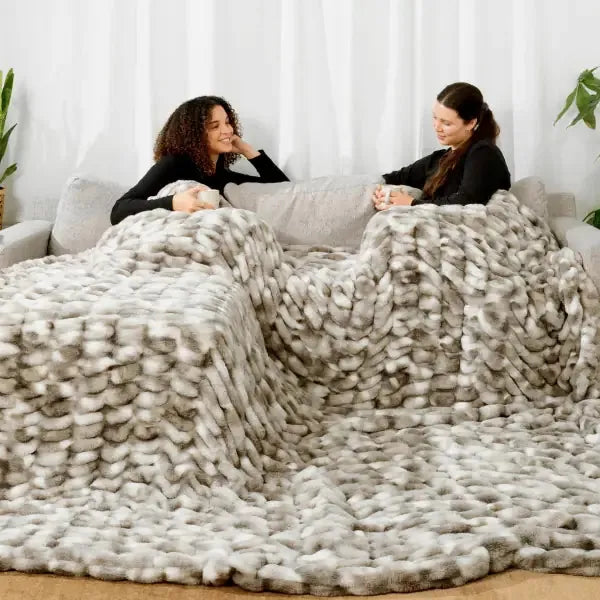 Two women sitting on a couch wrapped in a chunky knit Cloud Luxe Blanket in cream and gray tones